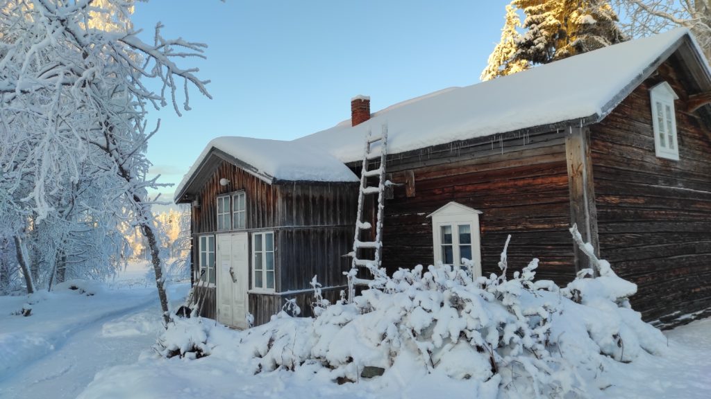 Fasade av brunbeiseit tømmerhus, dekket av snø.