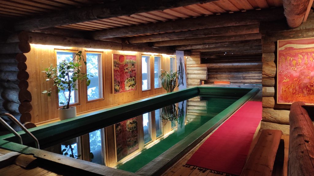 Basseng inne i et rom i et hus av tømmerstokker. Malerier i mange farger henger på veggene.
