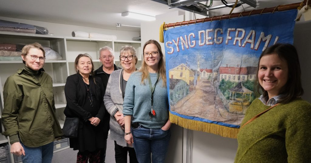 Seks personer står ved siden av en fane som har et maleri av et eldre bymiljø og teksten "Syng deg fram" over. Personene ser inn i kameraet og smiler.